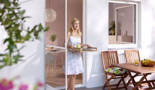 Schlanke blonde Frau in weissem Kleid trägt ein Tablett mit Essen und läuft dabei durch eine Tür mit Insektenschutzgitter nach draussen auf die Terrasse