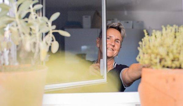 Frau mit kurzen blondierten Haaren steht innen am Fenster und öffnet ein Insektengitter Pendelfenster und lacht dabei. Aussen am Fenster stehen 2 Blumentöpfe mit Kräutern