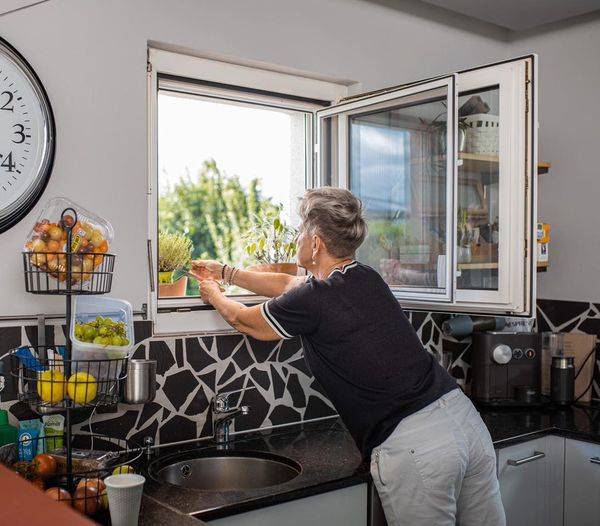 Frau steht innen am Fenster und hat das Pendelfenster mit Fliegengittergewebe geöffnet, um Kräuter in der Blumenkiste abzuschneiden